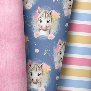 Könnte beinhalten: Ein blauer Stoff mit einem sich wiederholenden Muster aus weißen Comic-Pferden mit rosa Blumen und bunten Spritzern. Der Stoff liegt neben einem rosa Stoff und einem gestreiften Stoff mit rosa, gelben und blauen Streifen.