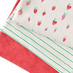 Peut inclure: Tissu plié avec une base rouge, une couche blanche à rayures vertes et une couche supérieure avec un motif de fraises. Les fraises sont rouges avec des tiges vertes, et les rayures sont espacées uniformément.