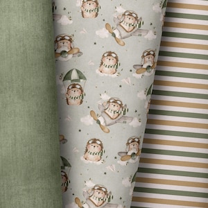 May include: Fabric rolls showing a repeating pattern of cartoon hedgehogs in aviator outfits, clouds, and paper aeroplanes on a pale green backdrop. Next to it is a plain green fabric and a striped design in green and beige.
