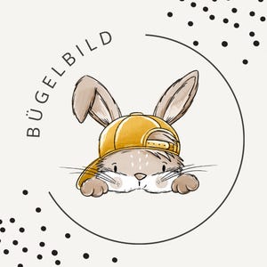Puede incluir: Ilustración de dibujos animados de un conejito marrón con una gorra de béisbol amarilla, asomándose por encima de una superficie. El conejito está dentro de un contorno circular negro con la palabra "BÜGELBILD". Puntos negros dispersos alrededor del círculo.