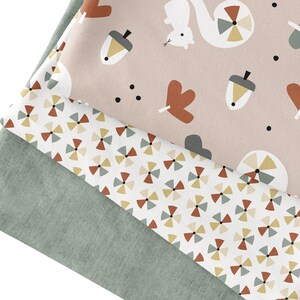 May include: Three folded fabrics with different patterns. The top fabric is light pink with a repeating pattern of acorns, leaves, squirrels, and dots. The middle fabric has a white background with a repeating geometric pattern. The bottom fabric is solid green.