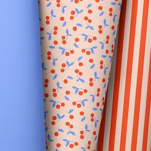 May include: A close-up of three rolled fabrics. One is solid light blue, the second has a pattern of red cherries with blue leaves on a beige background, and the third has vertical orange and white stripes.