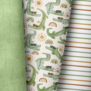 Op de afbeelding: Drie rollen stof met verschillende patronen. Eén rol is effen lichtgroen. De tweede rol heeft een herhalend patroon van groene alligators met zonnebrillen, regenbogen en zonnen op een crèmekleurige achtergrond. De derde rol heeft horizontale strepen in groen-, bruin- en crèmetinten.