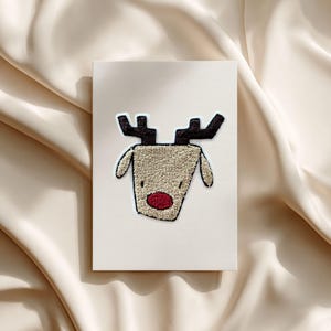 Könnte beinhalten: Eine cremefarbene Grußkarte mit einem Rentier-Design. Das Rentier hat einen strukturierten, beigefarbenen Körper, schwarze Geweihe und eine rote Nase. Die Karte ist rechteckig und liegt auf einer beigen, satinartigen Oberfläche.