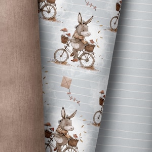 Op de afbeelding: Een grijs-wit gestreepte stof met een herhalend patroon van een ezel die op een fiets rijdt met een mand vol paddenstoelen. De ezel draagt een bruine sjaal en heeft een bruin-wit gestreepte staart. De achtergrond is lichtblauw met een witte vlieger.