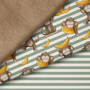Op de afbeelding: Stof met een patroon van apen en bananen. De bruine apen dragen zwart-witte bandana's en de bananen zijn geel. De achtergrond is wit met groene strepen. Een effen bruine stof is over de patroonstof gevouwen.
