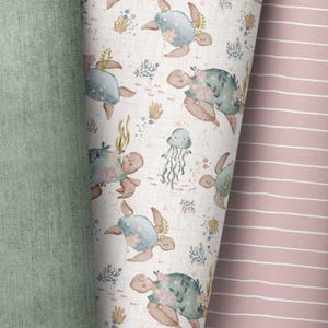 May include: A white fabric with a repeating pattern of blue and green sea turtles with pink flowers. The turtles are swimming in a light blue ocean with coral and seaweed. The fabric is bordered by a solid green fabric on the left and a pink and white striped fabric on the right.