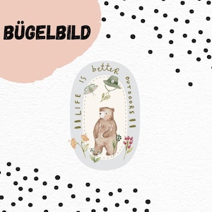 May include: An iron-on patch with the text "BÜGELBILD" in black, above an oval design. The oval design features a bear, a hat, a butterfly, and the words "Life is better outdoors". The background is white with black polka dots.