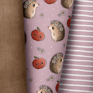 Op de afbeelding: Gerolde stof met een herhalend patroon van cartoon egels met madeliefkransen en appels met een bril op een roze achtergrond. De stof rechts heeft witte en roze strepen. Links is een bruine stof.