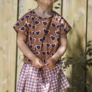 Puede incluir: Una persona joven que lleva una camisa de manga corta con estampado marr&oacute;n y rosa, una falda de cuadros rosa y blanca y un sombrero de paja marr&oacute;n.