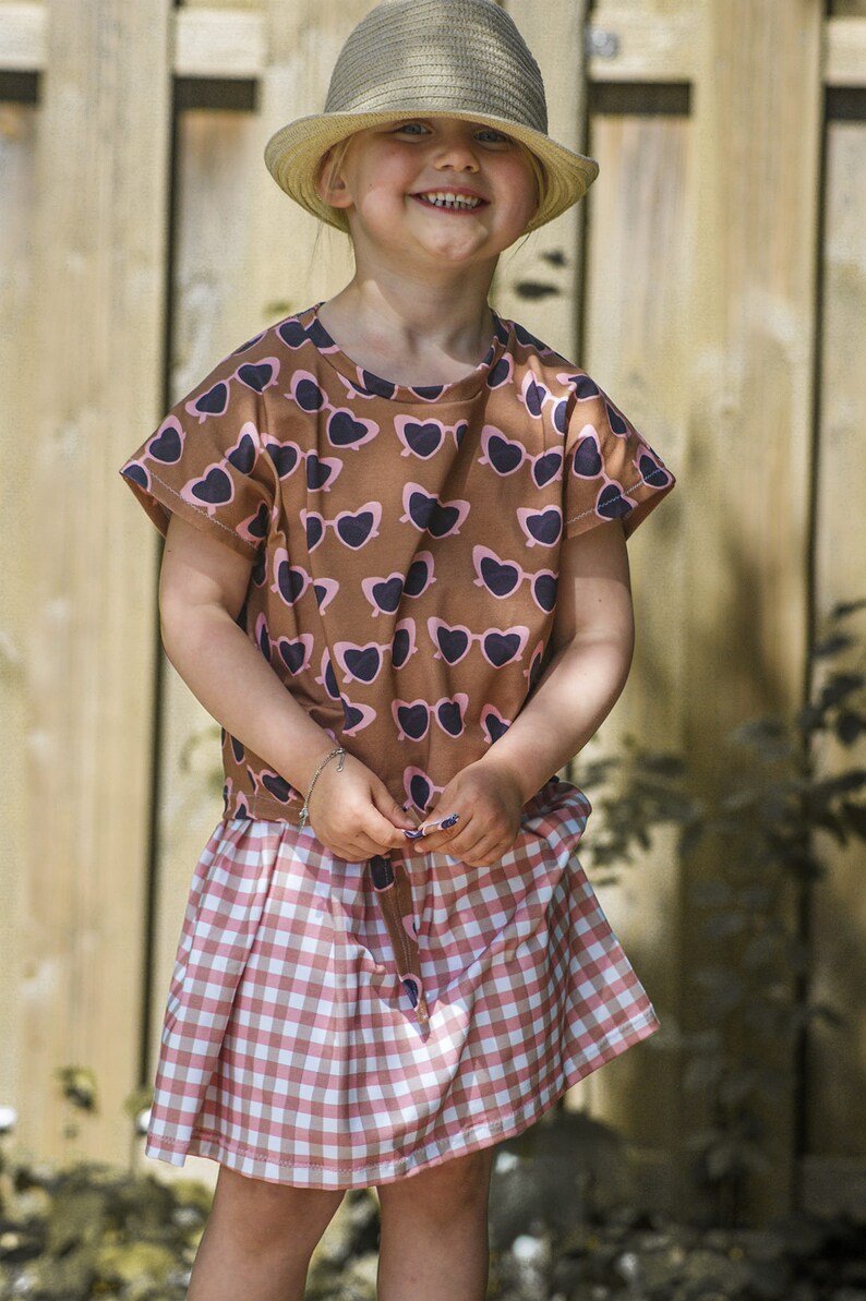 Puede incluir: Una persona joven que lleva una camisa de manga corta con estampado marr&oacute;n y rosa, una falda de cuadros rosa y blanca y un sombrero de paja marr&oacute;n.