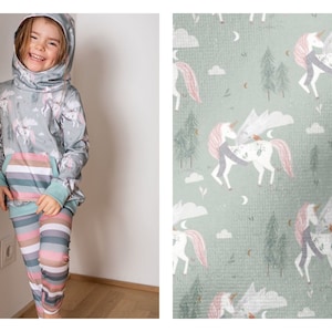 Könnte beinhalten: Ein Mädchen trägt ein graues Kapuzenpullover mit einem Einhorn-Druck und passende Leggings mit einem Regenbogen-Streifenmuster. Der Stoff hat ein sich wiederholendes Muster aus Einhörnern mit Flügeln und Wolken auf einem hellgrünen Hintergrund.