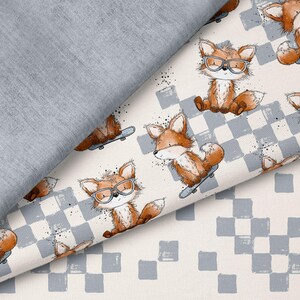 May include: Material featuring a repeating pattern of cartoon foxes on skateboards, wearing glasses. The foxes are brown and white, set against a cream background with a blue and white chequered pattern. A folded grey fabric is visible.