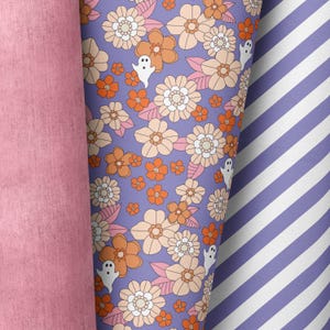 Könnte beinhalten: Lilafarbener Stoff mit einem sich wiederholenden Muster aus weißen und orangefarbenen Blumen und weißen Geistern. Der Stoff ist über einen rosa Stoff mit strukturierter Oberfläche gefaltet.