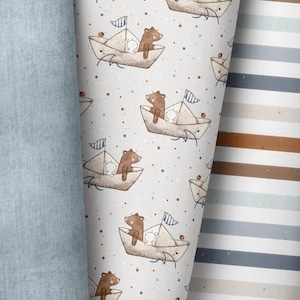 Könnte beinhalten: Nahaufnahme von drei verschiedenen Stoffmustern. Ein Stoff hat eine hellblaue Denim-Textur. Der zweite Stoff hat ein sich wiederholendes Muster aus einem Bären und einem Kaninchen in einem Papierboot. Der dritte Stoff hat horizontale Streifen in Braun- und Blautönen.
