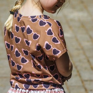 Puede incluir: Una ni&ntilde;a peque&ntilde;a con una camiseta de color marr&oacute;n y rosa con estampado de gafas de sol en forma de coraz&oacute;n y una falda de cuadros vichy blanca y rosa. Lleva un sombrero de paja y tiene el pelo rubio trenzado.