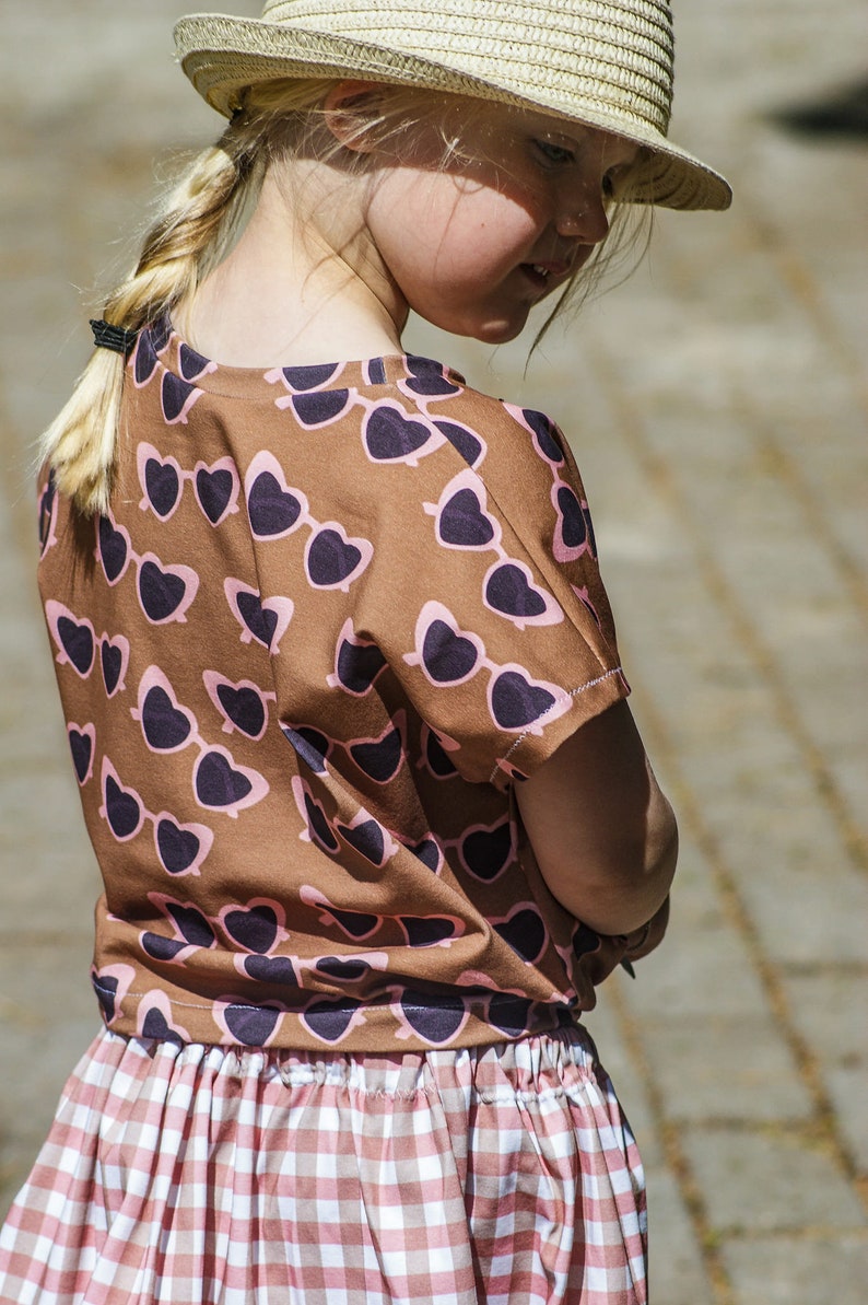 Puede incluir: Una ni&ntilde;a peque&ntilde;a con una camiseta de color marr&oacute;n y rosa con estampado de gafas de sol en forma de coraz&oacute;n y una falda de cuadros vichy blanca y rosa. Lleva un sombrero de paja y tiene el pelo rubio trenzado.