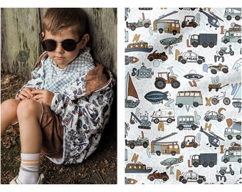 Puede incluir: Un niño pequeño con una chaqueta blanca y marrón con un patrón de vehículos y letras. La chaqueta tiene una cremallera y una capucha. El niño lleva gafas de sol y pantalones cortos marrones. El fondo es una valla de madera.