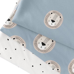 Könnte beinhalten: Nahaufnahme von Babybettwäsche mit hellblauem Hintergrund. Die Bettwäsche zeigt ein sich wiederholendes Muster von Löwengesichtern mit beigefarbenen Mähnen und schwarzen Details. Ein weißes Laken mit schwarzen Punkten ist ebenfalls zu sehen.
