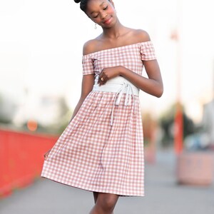 Puede incluir: Una joven con un vestido de hombros descubiertos de cuadros rosa y blanco con un cintur&oacute;n blanco. Est&aacute; de pie en una acera con una barandilla roja en el fondo.