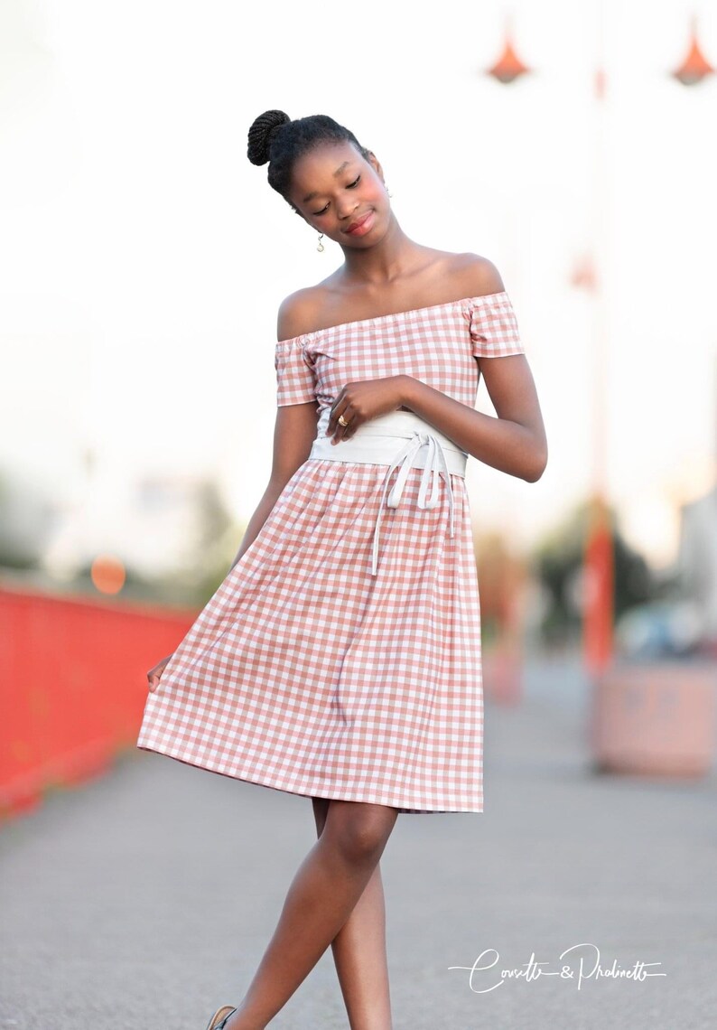 Puede incluir: Una joven con un vestido de hombros descubiertos de cuadros rosa y blanco con un cintur&oacute;n blanco. Est&aacute; de pie en una acera con una barandilla roja en el fondo.