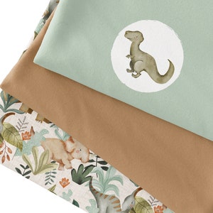 Op de afbeelding: Drie stofstalen met een dinosaurus thema. De bovenste staal is een lichtgroene stof met een witte cirkel met daarin een bruine dinosaurus. De middelste staal is een bruine stof. De onderste staal is een witte stof met een bruine dinosaurus, groene bladeren en oranje bloemen.