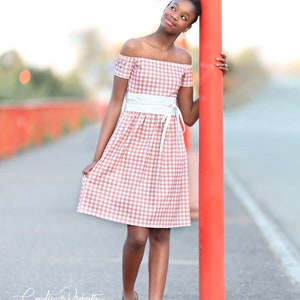 Puede incluir: Una persona joven lleva un vestido de hombros descubiertos de cuadros rosa y blanco con un cintur&oacute;n blanco. Est&aacute; de pie junto a un poste rojo y mirando hacia arriba.