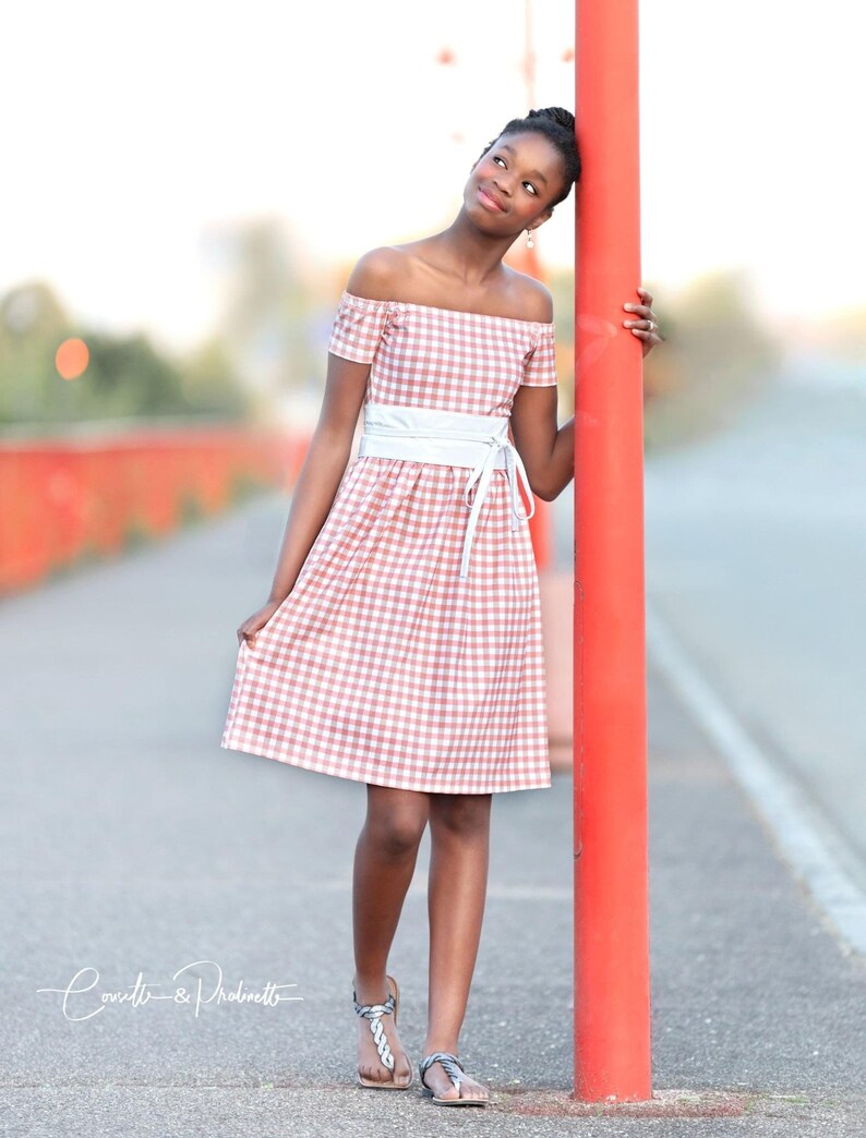 Puede incluir: Una persona joven lleva un vestido de hombros descubiertos de cuadros rosa y blanco con un cintur&oacute;n blanco. Est&aacute; de pie junto a un poste rojo y mirando hacia arriba.