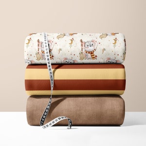 Peut inclure: Trois rouleaux de tissu empilés les uns sur les autres. Le rouleau du haut a un fond blanc avec un motif répété d'une chouette grise portant des lunettes et tenant une baguette magique. Le rouleau du milieu a un motif rayé jaune et marron. Le rouleau du bas est de couleur marron unie. Un mètre ruban blanc est enroulé autour des rouleaux.