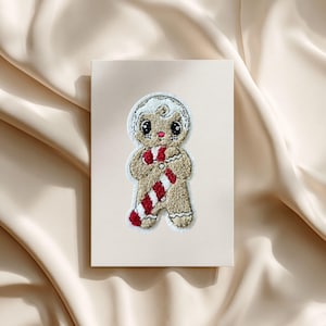 Könnte beinhalten: Ein skurriles Lebkuchenmann-Design auf einer Leinwand. Der Lebkuchenmann hat einen braunen Körper, weiße Zuckergussdetails und einen rot-weiß gestreiften Schal. Das Kunstwerk ist vor einem neutralen Hintergrund platziert.