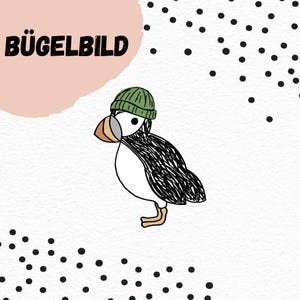 Può includere: Illustrazione di una pulcinella di mare con un berretto verde, con il testo tedesco "BÜGELBILD" sopra una forma rosa. La pulcinella di mare è nera, bianca e beige. Punti neri sparsi su sfondo bianco.