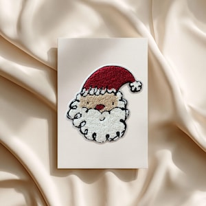 Könnte beinhalten: Eine Weihnachtskarte mit einem Weihnachtsmann-Design. Das Gesicht des Weihnachtsmanns hat eine rote Mütze, ein beiges Gesicht, einen weißen Bart und eine rote Nase. Die rechteckige Karte hat einen cremefarbenen Hintergrund. Das Weihnachtsmann-Design besteht aus strukturiertem Stoff.