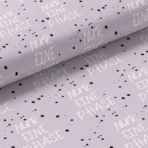 Könnte beinhalten: Helllila Stoff mit dem weißen Schriftzug "NUR EINE PHASE" und schwarzen Tintenspritzern. Der Stoff ist gefaltet, wodurch seine Textur und sein Muster sichtbar werden. Der Satz bedeutet "nur eine Phase".