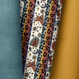 Puede incluir: Una tela blanca con un patrón repetido de rayas de colores, estampado de leopardo e ilustraciones de estilo retro. Las ilustraciones incluyen estrellas, arcoíris, lunas, signos de paz y rayos.