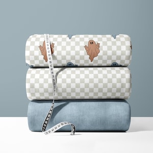 Könnte beinhalten: Drei Stoffrollen mit einem karierten Muster und einem Geisterdesign. Der Stoff ist beige mit einem hellgrauen Karomuster und braunen Geistern. Die oberste Rolle hat ein blaues Karomuster mit grauen Geistern. Ein weißes Maßband ist um die Rollen gewickelt.