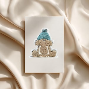 Puede incluir: Tarjeta de felicitación con un elefante de dibujos animados que lleva un gorro de punto azul con un pompón. El elefante es marrón con colmillos blancos y está sentado. La tarjeta está sobre un fondo crema.