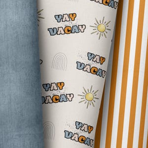Op de afbeelding: Gerolde stof met een herhaald patroon van de woorden "YAY VACAY" in blauw en oranje, samen met illustraties van zon en regenboog. De stof is crèmekleurig met zwarte spikkels. Denim stof en een gestreepte stof zijn zichtbaar.