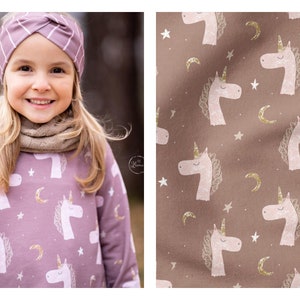 May include: A young girl wearing a purple long-sleeved dress with a unicorn print and a pink headband. The dress has a pattern of white unicorns, gold stars, and crescent moons. The background is a brown fabric with a similar unicorn print.