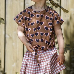 Puede incluir: Una ni&ntilde;a peque&ntilde;a con una camiseta marr&oacute;n y rosa con un estampado de gafas de sol en forma de coraz&oacute;n y una falda de cuadros rosa y blanca. Lleva un sombrero de paja y tiene el pelo recogido en una coleta.