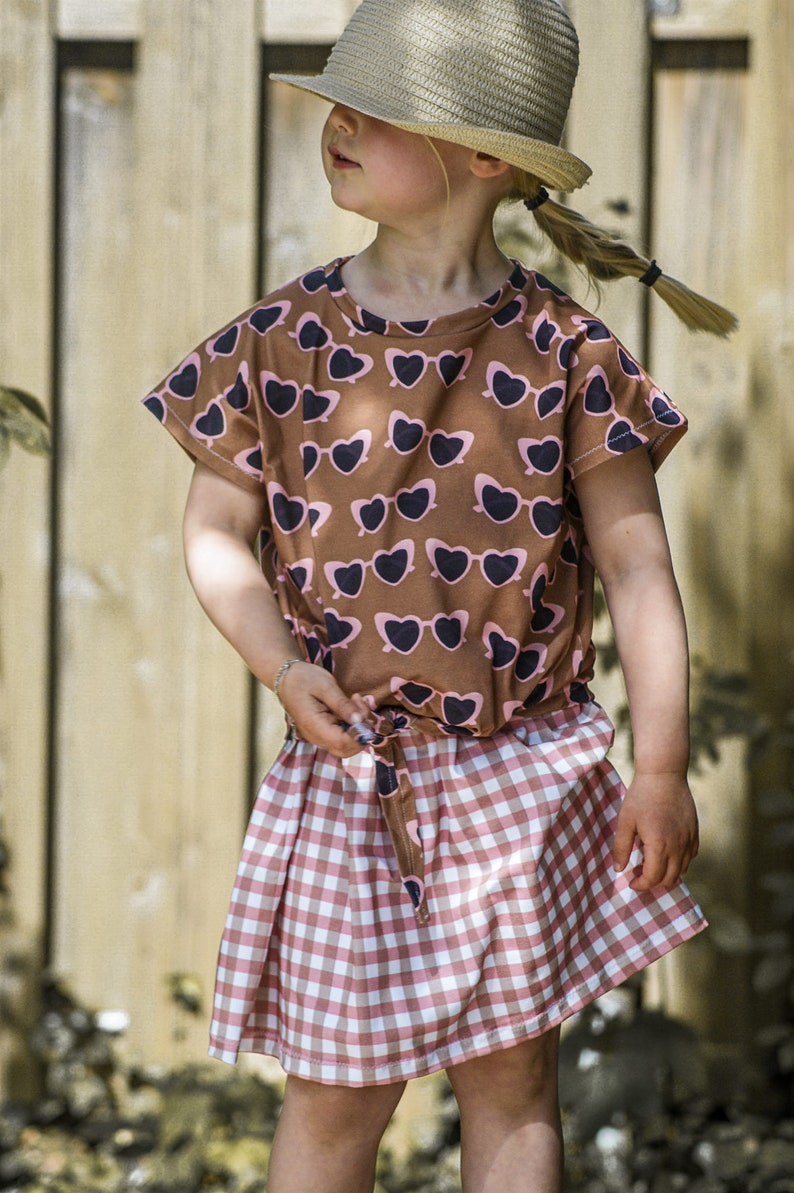 Puede incluir: Una ni&ntilde;a peque&ntilde;a con una camiseta marr&oacute;n y rosa con un estampado de gafas de sol en forma de coraz&oacute;n y una falda de cuadros rosa y blanca. Lleva un sombrero de paja y tiene el pelo recogido en una coleta.