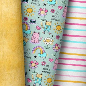 Könnte beinhalten: Nahaufnahme von drei verschiedenen Stoffmustern. Eines ist einfarbig gelb, das zweite hellblau mit einem sich wiederholenden Muster aus Katzen, Blumen, Regenbogen und dem Text "MY WORLD IS WONDERFUL". Das dritte ist weiß mit rosa, gelben und blauen Streifen.