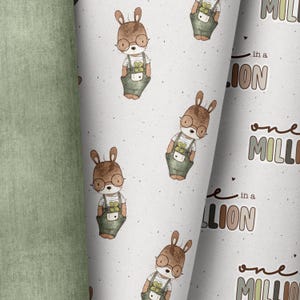 Op de afbeelding: Een witte stof met een herhalend patroon van bruine konijnen die een bril en groene overalls dragen. De tekst "one in a million" wordt herhaald in bruine, groene en beige letters.