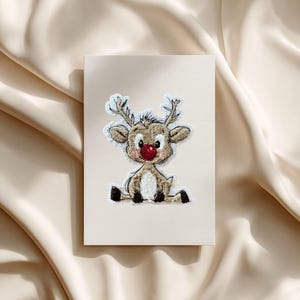 Könnte beinhalten: Eine beige Grußkarte mit einem gestickten Rentier-Design. Das Rentier hat einen braunen Körper, eine rote Nase und weiße Geweihe. Die Karte ist vor einem beigen, strukturierten Hintergrund platziert und erzeugt eine weiche, festliche Ästhetik.