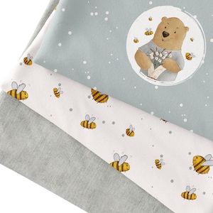 Könnte beinhalten: Drei Stoffbahnen mit weißem Hintergrund und einem sich wiederholenden Muster aus gelben und schwarzen Bienen. Die obere Bahn ist hellblau mit weißen Punkten und einem Kreis, in dem ein brauner Bär einen Strauß weißer Blumen hält. Der Bär trägt ein hellblaues Hemd.