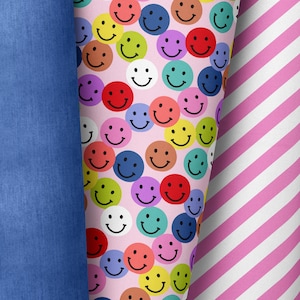 Può includere: Un tessuto rosa con un motivo ripetuto di facce sorridenti colorate. Il tessuto è mostrato accanto a un tessuto denim blu e un tessuto a righe rosa e bianco.