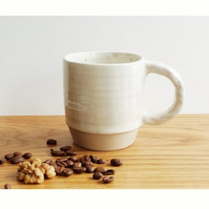 Può includere: Una tazza in ceramica bianca con un manico testurizzato e una glassa leggermente avorio. La tazza è appoggiata su una superficie di legno con chicchi di caffè e noci sparsi intorno.
