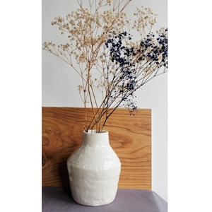 Puede incluir: Jarrón de cerámica blanco con superficie texturizada, lleno de flores secas blancas y azules.