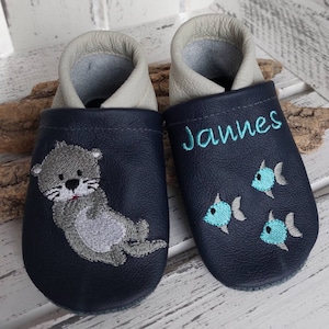Könnte beinhalten: Ein Paar dunkelblaue Babyschuhe mit einem grauen Seeotter und türkisfarbenen Fischen, die darauf gestickt sind. Der rechte Schuh hat den Namen "Jannes" gestickt.