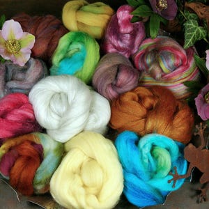 Könnte beinhalten: Eine Sammlung farbenfroher Wollvliese in Weiß, Gelb, Grün, Blau, Rosa und Braun. Die Wolle ist dekorativ mit Blumen und einer Metallfroschfigur angeordnet.