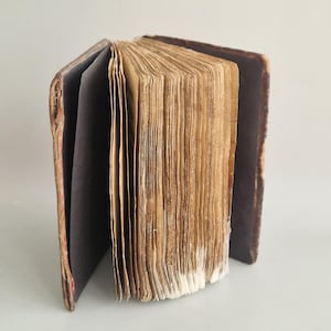 Pode incluir: Um livro antigo aberto com páginas amareladas e envelhecidas. A lombada e a capa do livro são marrons escuros, com sinais de desgaste. As páginas estão empilhadas, mostrando a textura do papel. As bordas são ásperas e irregulares.